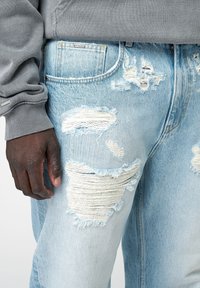 Vaqueros de mezclilla azul claro con grandes desgarros deshilachados, dobladillos sin rematar y una textura suave. Combinados con un suéter gris con puños acanalados.