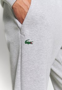 A kezében a világosszürke melegítőnadrág zsebében, amelynek a combján egy kis zöld krokodil-logó található.