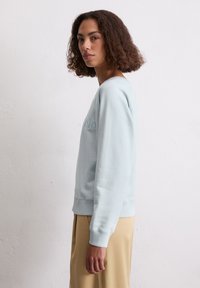 Młoda kobieta z kręconymi włosami, ubrana w jasnoniebieską bluzę i beżowe spodnie, stoi bokiem przy gładkiej białej ścianie.