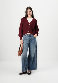 Bordeaux gebreide cardigan met een V-hals en drie knopen, gecombineerd met een wijd uitlopende blauwe jeans en zwarte ballerina's. Inclusief een bruine crossbodytas.