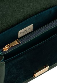 Grön läderhandväska med guld detaljer, som har en dragkedjor ficka och ett textilinteriör. Varumärkesetiketten "FABLE" är synlig inuti.