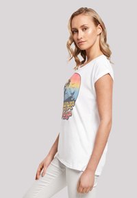 Weißes Baumwoll-T-Shirt mit kurzen aufgerollten Ärmeln, versehen mit einem bunten Retro-Sonnenuntergang-Graphic, Palmen und Text in der Mitte.