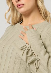 Jasnozielony sweter prążkowany z okrągłym dekoltem i przedłużonymi rękawami z detalem w postaci wiązania przy mankietach. Miękka tekstura dzianiny.