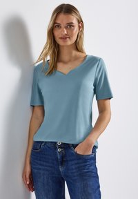 Ljusblå, kortärmad topp med V-ringning, gjord av mjuk tyg. Bärs tillsammans med mörkblå jeans som har en framficka och knapppdetaljer.