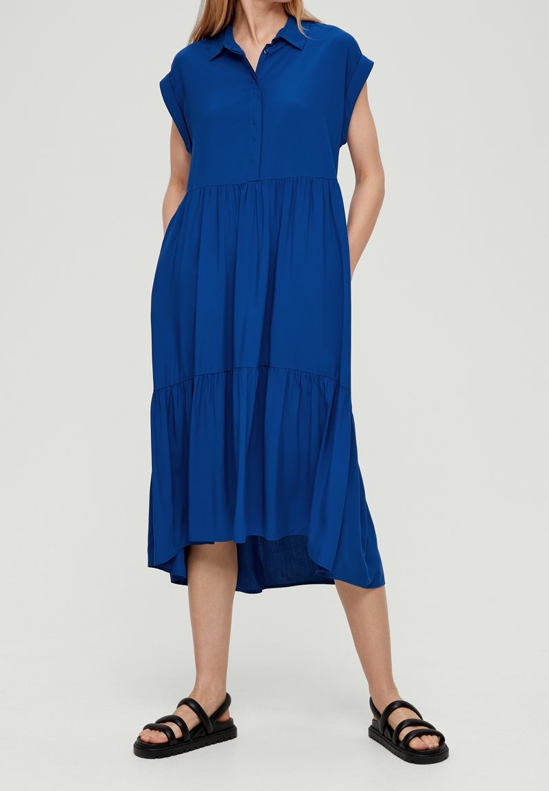 Femme portant une robe midi bleue à manches courtes avec col et des sandales plates noires, debout les mains dans les poches, devant un fond uni.
