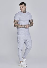 Camiseta ajustada gris claro y pantalones deportivos de una tela suave. Los pantalones deportivos cuentan con bolsillos laterales y puños acanalados. Zapatillas blancas con detalles en negro.