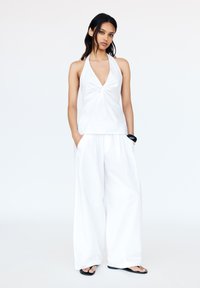 Top bianco senza maniche con dettaglio frontale attorcigliato, abbinato a pantaloni bianchi morbidi. Sandali neri sottili. Design minimalista su uno sfondo chiaro.