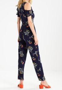Granatowy kombinezon w kwiaty z odkrytymi ramionami, falbaniastymi rękawami i prostymi nogawkami. Charakteryzuje się kolorowymi wzorami kwiatowymi na całej powierzchni.