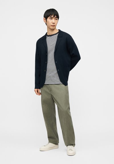 Jeune homme debout portant un cardigan noir sur une chemise à rayures noires et blanches, un pantalon olive et des baskets blanches sur un fond uni.