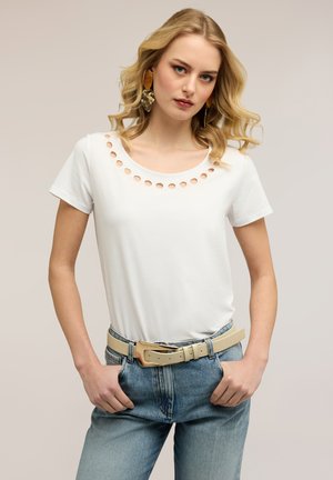 Blonde woman with wavy hair wearing white short-sleeve top with circular cutouts at neckline, blue jeans, beige belt, and large earrings.