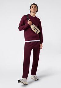 Set di felpa e pantaloni bordeaux, in misto cotone, dal taglio comodo. Completato da una piccola borsa a vita beige con zip, indossata crossbody.