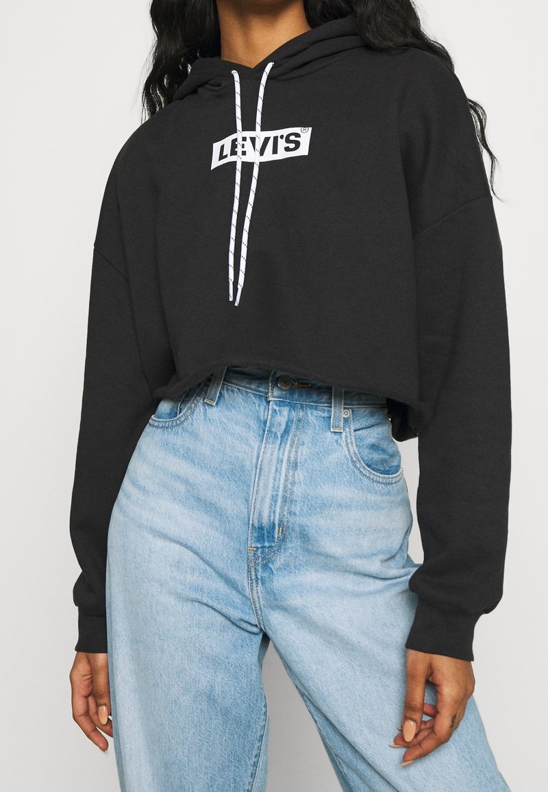 Personne portant un sweat à capuche court noir Levi’s avec des cordons blancs et un jean taille haute bleu clair.