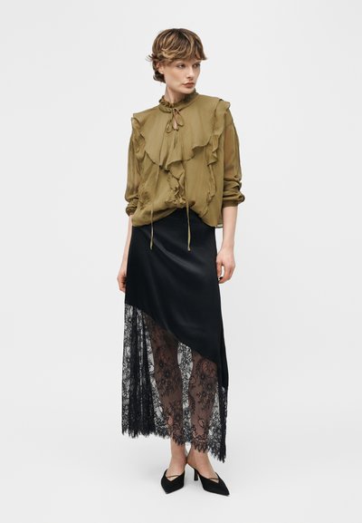Γυναίκα με κοντά μαλλιά, που φορεί μια ελιά πουκάμισο με βολάν και μαύρη ασύμμετρη φούστα με διάφανη δαντέλα επικάλυψη και μαύρες μυτερές γόβες.