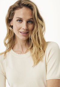 Mujer rubia con ojos azules sonriendo suavemente, vistiendo una blusa corta de manga corta color crema, contra un fondo blanco.