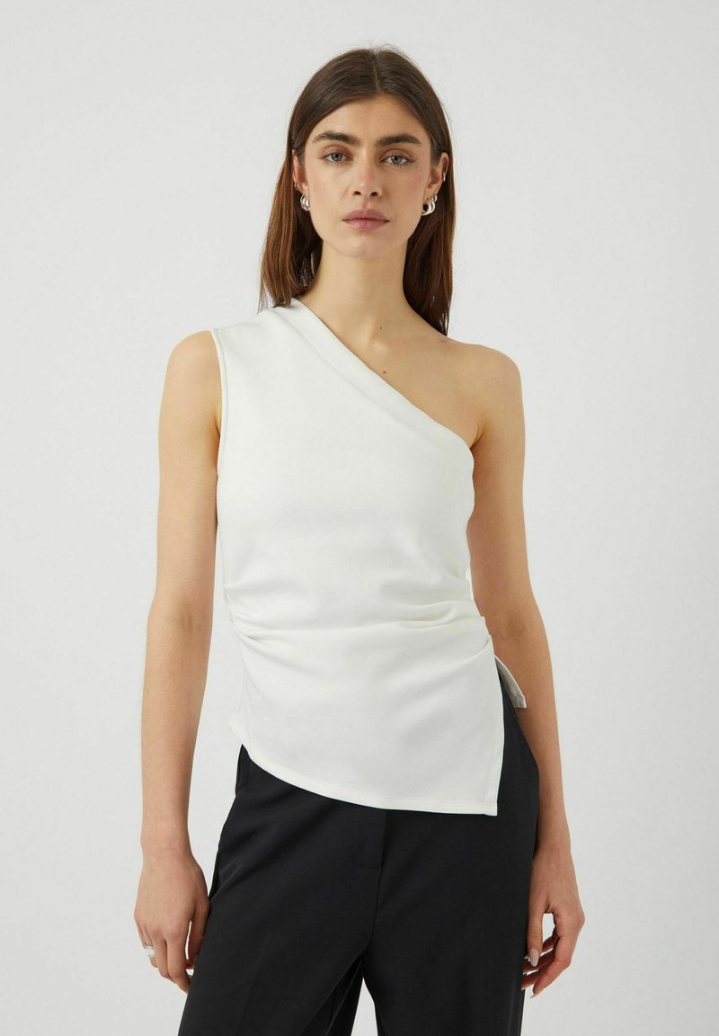 Top blanco de un solo hombro en tejido suave, con detalle fruncido en el costado, dobladillo asimétrico y silueta elegante. Combinado con pantalones negros.