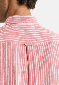 Camicia a righe rosse e bianche con colletto button-down, caratterizzata da un dettaglio a yoke sul retro e una piega per una maggiore libertà di movimento.
