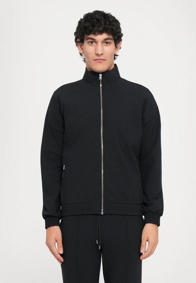 Sort zip-jakke med ståkrave, strækbare manchetter og kant. Glat stof med en tætsiddende design, med sidelommer og en metallisk lynlås.
