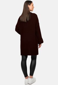 Maglione lavorato a maglia di colore bordeaux scuro, lungo e dalla vestibilità ampia con trama a coste, caratterizzato da uno spacco centrale sul retro, abbinato a leggings neri e scarpe da ginnastica bianche.