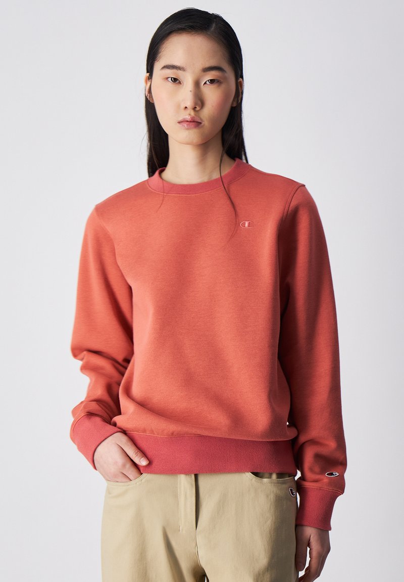 Sudadera coral de manga larga con puños y dobladillo acanalados. Presenta un pequeño logotipo bordado en el pecho y una textura suave.