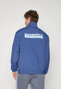 Blå lettvektsjakke med høy krage, med "NASHVILLE"-grafikk i lyseblått på ryggen, elastisk nederkant og mansjetter.