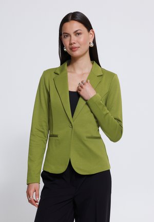 Vrouw met lang donker haar, met een groene blazer over een zwarte top en zwarte broek, poserend met de hand bij de kraag en een neutrale achtergrond.