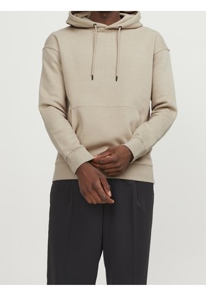 Un hoodie beige en mélange de coton, doté d'une poche kangourou, d'une capuche avec cordon de serrage ajustable et de poignets et ourlet côtelés. Porté avec un pantalon sombre.