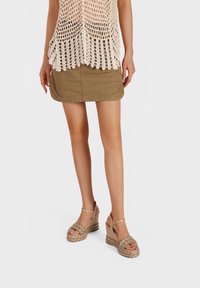 Top in crochet beige con orlo frangiato abbinato a una mini gonna khaki e sandali con zeppa color testa di moro caratterizzati da dettagli intrecciati e punta aperta.