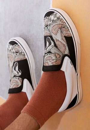Pieds portant des chaussures Vans blanches slip-on avec des motifs serpent et damier, associées à des chaussettes couleur rouille sur un fond gris et orange.