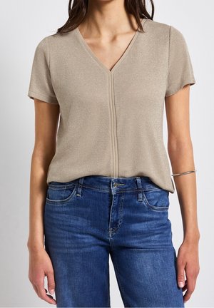 Kobieta ubrana w beżową błyszczącą bluzkę z krótkim rękawem i dekoltem w serek, zestawioną ze średnio jasnymi niebieskimi dżinsami oraz srebrną bransoletką na lewej ręce.