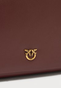 Μπορντό δερμάτινη τσάντα με χρυσό κυκλικό μεταλλικό διακοσμητικό με δύο πουλιά και ανάγλυφο όνομα μάρκας, λεία υφή και δομημένος σχεδιασμός.