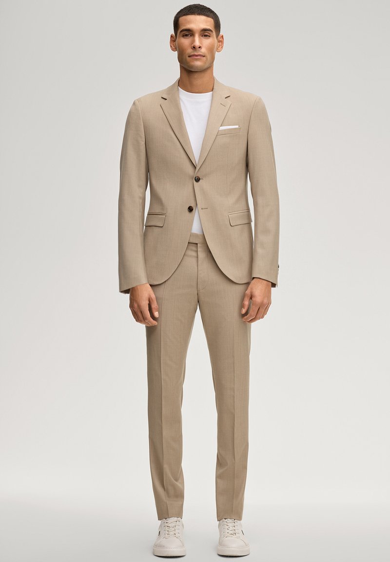 Mann in beigem Slim-Fit-Anzug mit weißem T-Shirt und weißen Turnschuhen, stehend vor einfachem hellem Hintergrund.