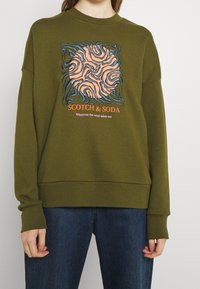 Oliwkowy sweatshirt z okrągłym dekoltem, z wielokolorowym abstrakcyjnym wzorem i napisem "SCOTCH & SODA" na środku.