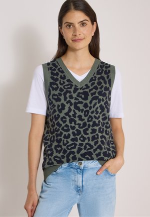 Frau trägt eine grüne und schwarze ärmellose Pulloverweste mit Leopardenmuster über einem weißen T-Shirt und hellblauen Jeans, steht mit einer Hand in der Tasche.