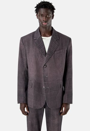 Uomo che indossa una giacca da completo oversize a quadri grigio scuro con pantaloni abbinati sopra una camicia bianca, in piedi su uno sfondo semplice.