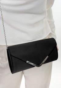 Black clutch bag made of smooth material, featuring a sleek, triangular flap and a silver chain strap, with a metallic accent on the front.
