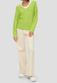 Grüner gestrickter V-Ausschnitt-Pullover kombiniert mit cremefarbenen, ausgestellten Jeans und weißen Plateausneakern. Lässige, lockere Passform mit geripptem Struktur.