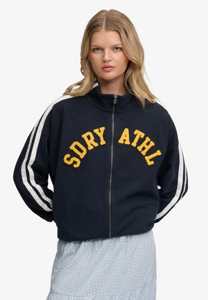 Młoda kobieta o blond włosach nosi granatową kurtkę z białymi paskami na rękawach i żółtymi literami „SDRY ATHL” z przodu, w połączeniu z jasnoniebieską spódnicą.