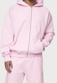 Felpa rosa con zip realizzata in un tessuto morbido, dotata di cappuccio, polsini e fondo a coste, e tasche frontali. Branding discreto sul petto.