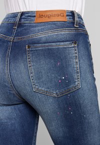 Blå denimjeans med en läderlogotyp, märkbara färgfläckar i rosa och vitt, samt traditionell sömnad och fickor.