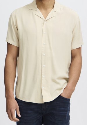 Mand iført beige kortærmet skjorte med knapper og krave, med den ene hånd i lommen på mørkeblå jeans, mod en ensfarvet lys baggrund.