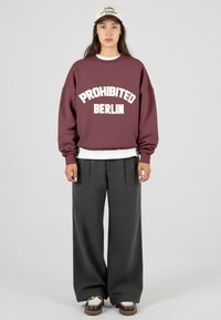 Žena v bílé čepici, vínovém svetru s nápisem "PROHIBIT BERLÍN", bílém tričku pod ním, širokých tmavě šedých kalhotách a hnědých platformových botách.