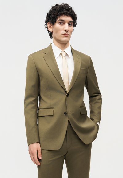 Άνδρας με κατσαρά μαλλιά που φοράει πράσινο-ελιά κοστούμι, λευκό πουκάμισο και μπεζ γραβάτα, στέκεται με το ένα χέρι στη τσέπη του μπροστά σε απλό φόντο.