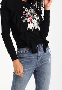 Un pull noir avec des détails en volants, orné de broderies florales rouges et blanches. Associé à un jean bleu déchiré, affichant un look décontracté.