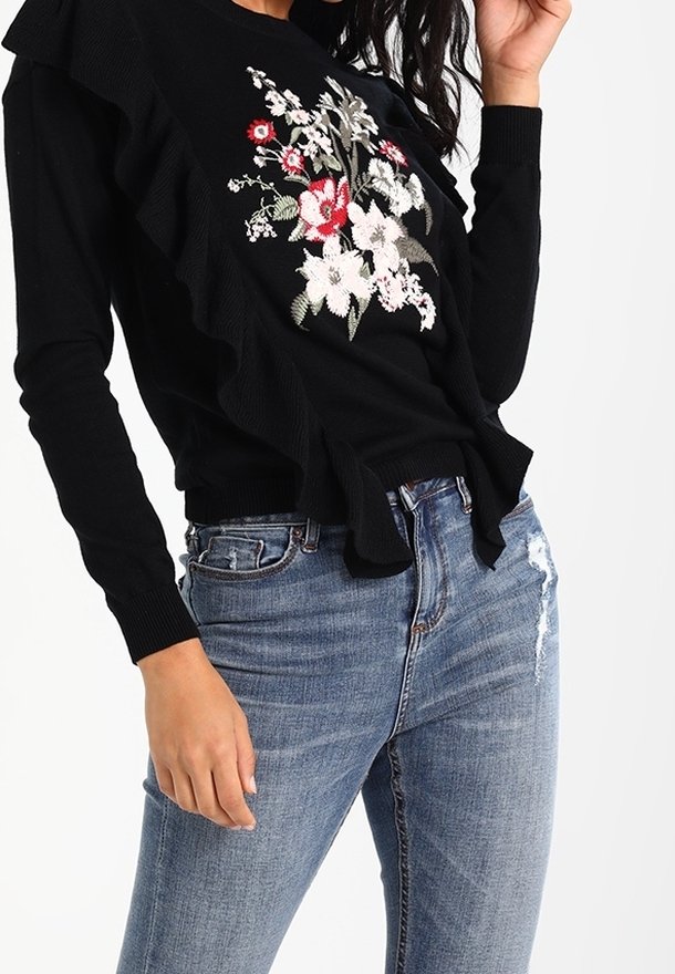 Un pull noir avec des détails en volants, orné de broderies florales rouges et blanches. Associé à un jean bleu déchiré, affichant un look décontracté.