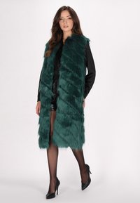 Langer Kunstfell-Westen in tiefem Teal, mit einem Zickzackmuster und einem kragenlosen Design. Getragen über einem schwarzen Oberteil und glänzenden Shorts.