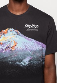 Svart bomullströja med en färgglad berggrafik i nyanser av orange, lila och blå, samt texten "Sky High Adventures".