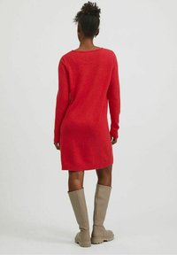 Robe en maille rouge à coupe décontractée avec un ourlet côtelé, associée à des bottes beige montantes jusqu'aux genoux. La robe présente des manches longues et un col rond.