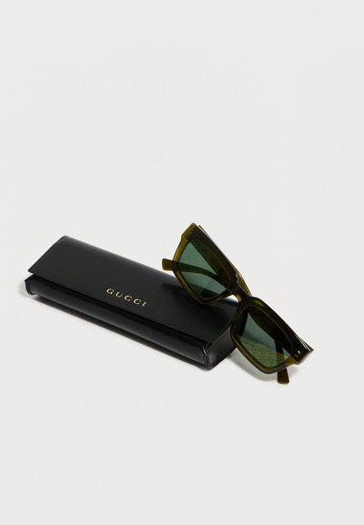 Lunettes de soleil carrées teintées de vert reposant sur un étui rectangulaire noir Gucci avec des lettres dorées sur un fond blanc.