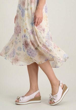 Kobieta w kwiecistej spódnicy midi i beżowych sandałach na koturnie z kokardką, stojąca na jednolitym jasnym tle.