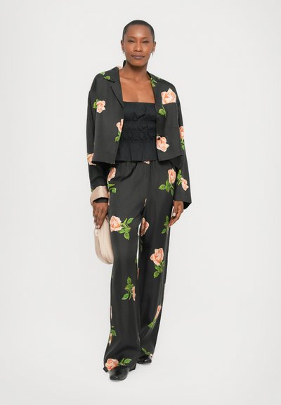 Costum floral negru cu modele de trandafiri roz, jachetă scurtă și pantaloni evazați. Modelul ține o poșetă bej deschis. Textură de material neted.